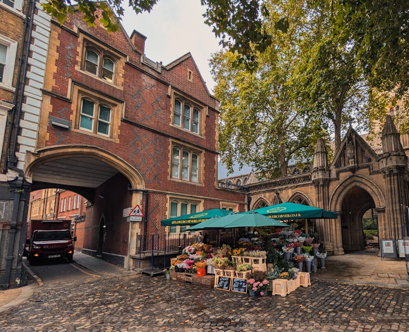 A bustling outdoor flower market set up on a cobbled street near a historic brick archway and gothic-style stone structures, with a large green canopy sheltering various colorful floral arrangements.