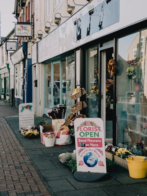 View of the exterior of a flower shop with large glass windows, displaying floral arrangements and decorations inside. Outside, on the sidewalk, various flower arrangements and supplies are arranged,